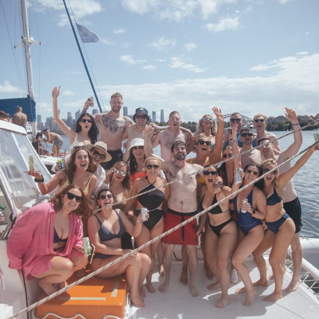 A group of people in swimwear are posing for a photo on a sailboat during a Sydney boat party hire. Some hold drinks, and they are smiling and waving at the camera. The background shows water and the city skyline.