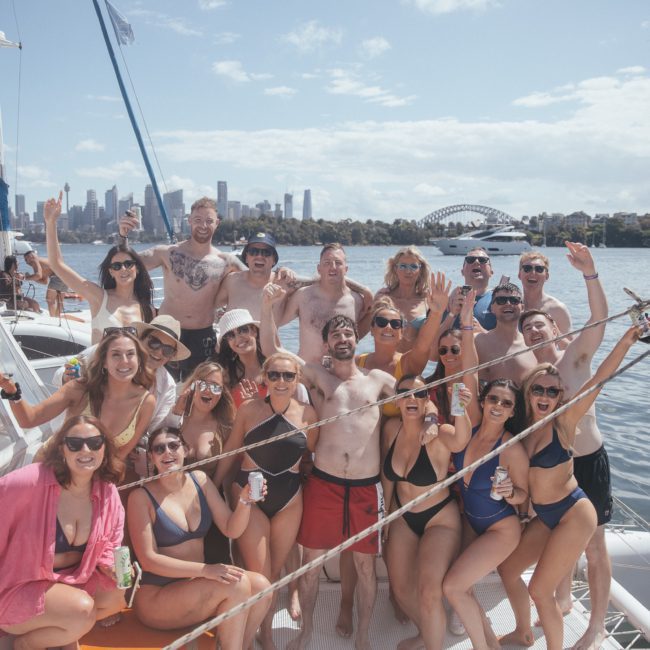 A group of people posing on a sailing boat in swimwear with a city skyline and a large bridge in the background. The weather appears sunny, and they seem to be celebrating or having a party, showcasing the excitement of Private yacht charter Sydney Harbour.