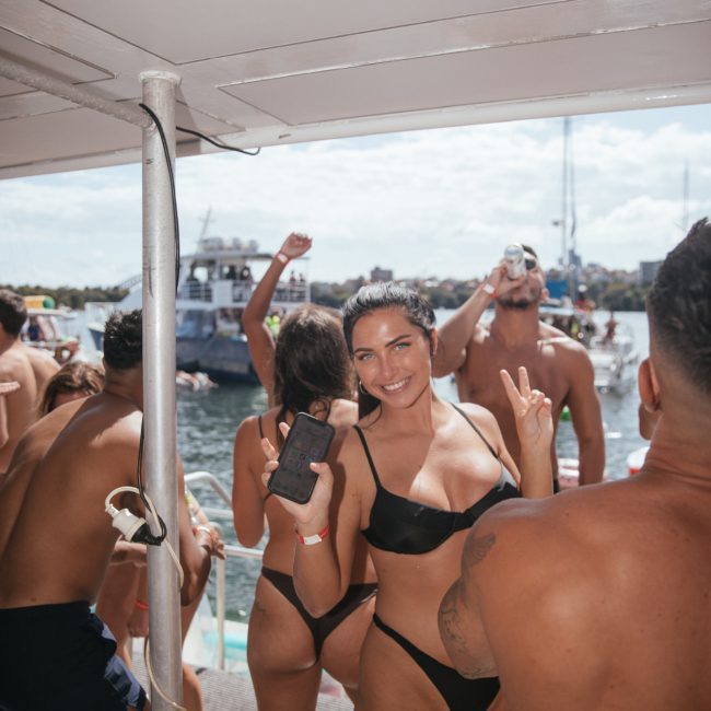 People in swimwear on a boat enjoying a sunny day, with one person at the forefront smiling and holding up a phone and a peace sign. Other boats are visible in the background on the water, setting a perfect scene for Sydney boat party hire.