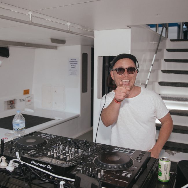 A man wearing sunglasses and a hat points at the camera while standing behind DJ equipment on a boat, perfect for a Sydney boat party hire.