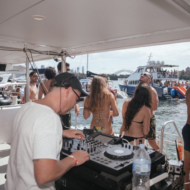 A DJ performs on a luxury yacht during a lively event. People in swimsuits are dancing and socializing, with other boats and a city skyline visible in the background.
