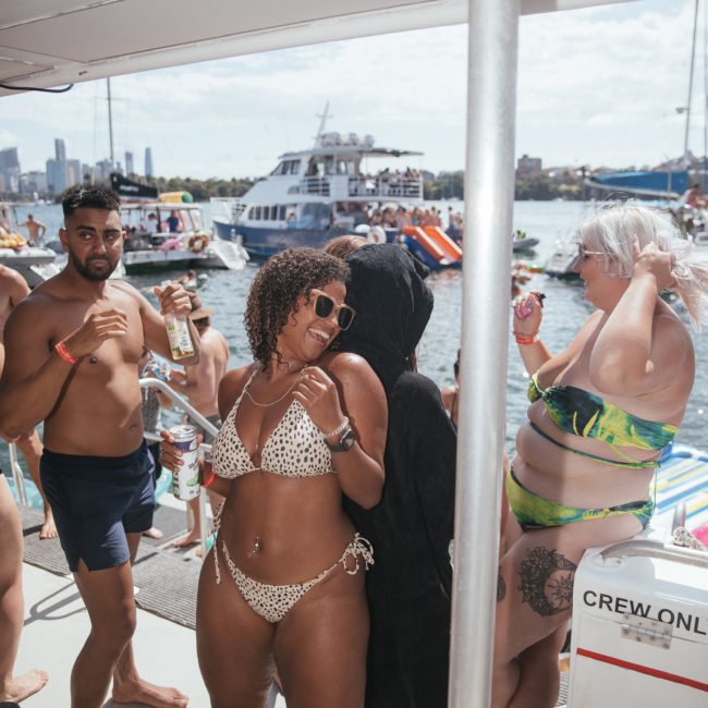 A group of people on a boat party, with several individuals wearing swimwear. Various boats are seen in the background on the water, including an elegant private yacht charter in Sydney Harbour.