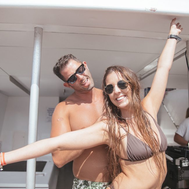 A man and woman, both wearing sunglasses, stand on a catamaran. The woman, in a bikini, has her arms outstretched and smiles at the camera while the shirtless man behind her also smiles. Others are in the background enjoying what appears to be a lively catamaran party in Sydney.