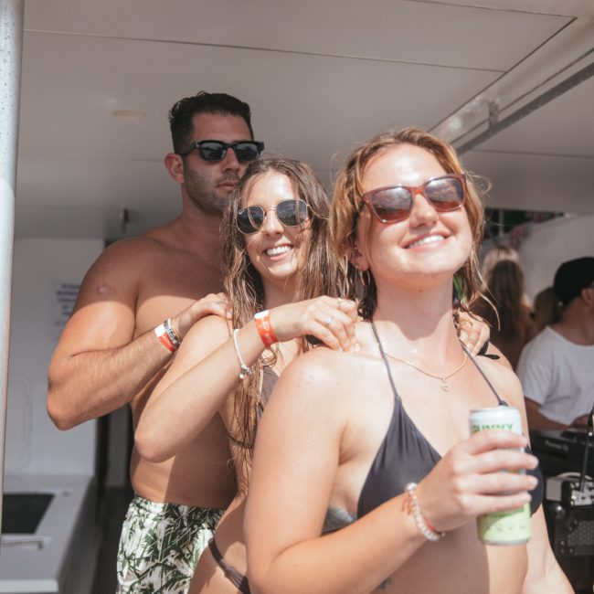 Three people in swimsuits and sunglasses stand closely on a boat, smiling and enjoying a drink. One is holding a can, another has a hair tie, and the third is standing behind them. Perfect moments like these are why Sydney boat party hire is so popular for unforgettable gatherings.