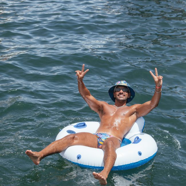 A person in a hat and swim trunks is floating on a white inflatable ring in the water, smiling, and making peace signs with both hands during a private yacht charter on Sydney Harbour.