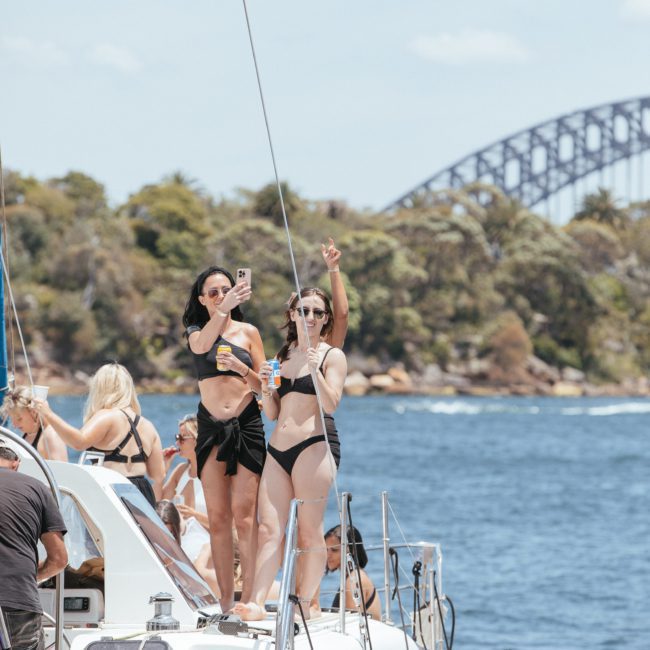 A group of people in swimsuits are standing on a luxurious yacht with two women in the foreground taking photos and holding drinks. The background features a bridge, trees, and a clear sky, perfect for a Sydney boat party hire.