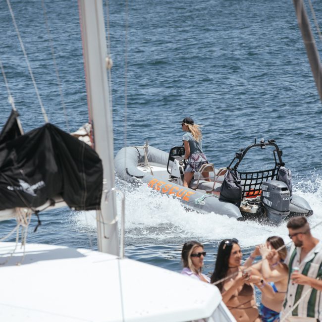 A woman in a small boat steers through the water while a group of people enjoy a Sydney boat party hire on a docked boat in the foreground.