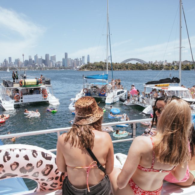 People on boats and inflatables enjoy a sunny day on the water, with a city skyline and bridge visible in the background, perfect for those considering Sydney boat party hire.
