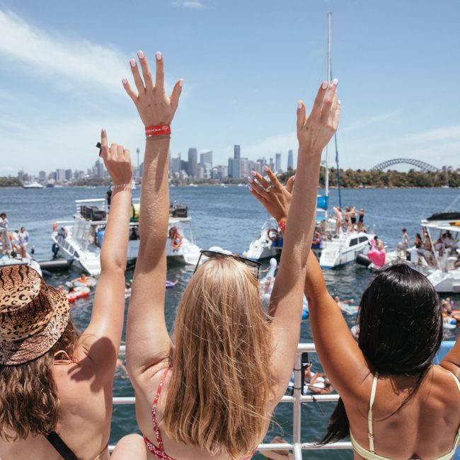 Three people stand on a boat with their arms raised, facing a crowded harbor with multiple boats and the city skyline in the background on a sunny day, enjoying what appears to be an exciting Sydney boat party hire event.