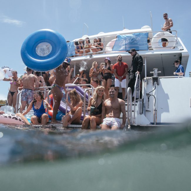 A large group of people enjoys a sunny day on a luxury yacht hire in Sydney, with some in swimsuits holding inflatable pool toys while others observe from the upper deck.