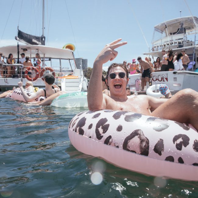 A man relaxes on a leopard-print inflatable in the water, making a peace sign. Several people are on nearby boats in the background, enjoying a sunny day. It looks like they're gearing up for an exciting Sydney boat party hire experience.