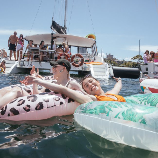 People enjoying a sunny day on inflatable floaties in the water, surrounded by boats and other people. A DJ boat hire Sydney adds an energetic vibe to the scene, making it the perfect spot for a Sydney boat party hire.
