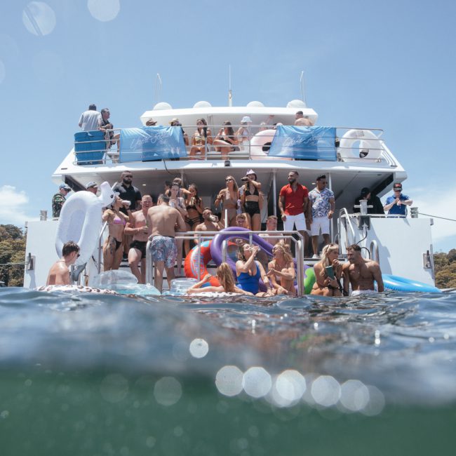 A large group of people are enjoying a Sydney boat party hire on a boat and in the water. Some are on the deck, while others are swimming or floating on inflatables. The sky is clear and the surroundings appear natural, making it perfect for a memorable event.