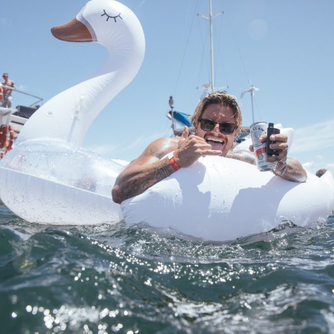 A person on an inflatable swan in the water holds a drink and gives a thumbs up, with boats and others in the background, enjoying a private yacht charter Sydney Harbour.