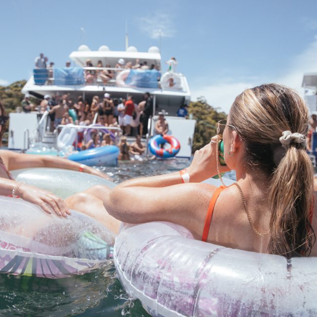 People relax on inflatable floats in the water while others gather on a boat in the background under a clear sky, enjoying their private yacht charter in Sydney Harbour.