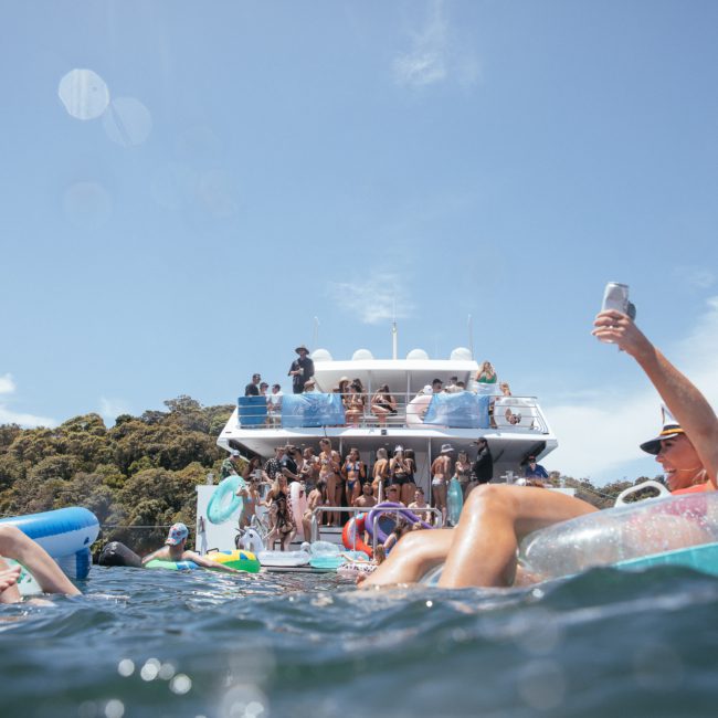 People on various inflatables relaxing in the water near boats with a large crowd gathered on a private yacht charter Sydney Harbour in the background under a clear blue sky.