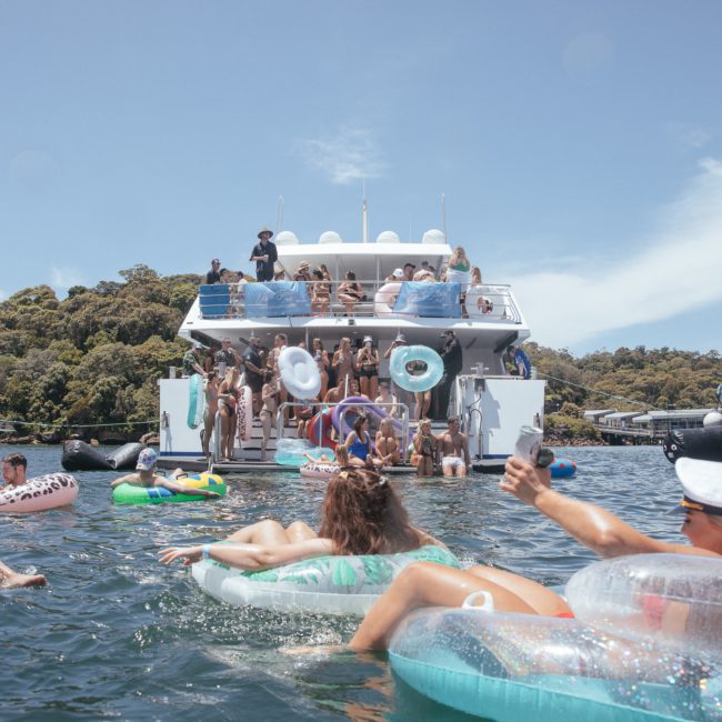 People are swimming and lounging on inflatable tubes in the water near a large boat. Several individuals are on the boat, enjoying a vibrant Sydney boat party hire, and other boats are visible in the background.