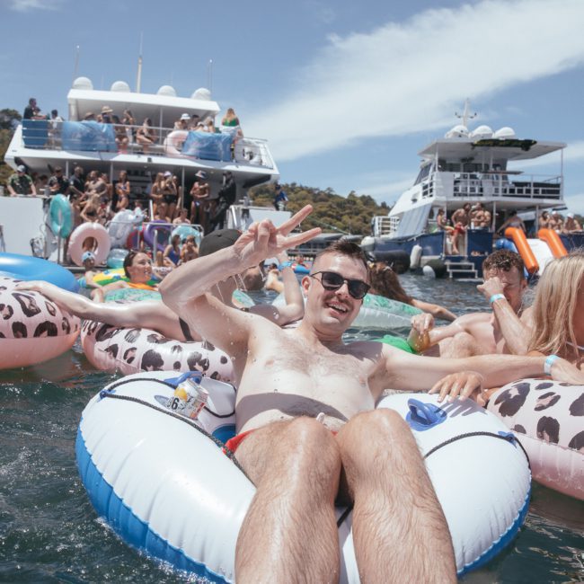 People are floating on inflatable pool rings in the water near boats on a sunny day. One man in the foreground is giving a peace sign while holding a drink, with the sleek lines of a luxury yacht hire Sydney framing the scene.