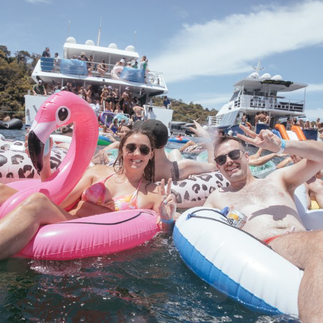 Two people float on inflatable rafts, with a woman on a pink flamingo and a man on a blue and white ring, both smiling and showing peace signs. Boats, including options for private yacht charters around Sydney Harbour, are in the background.