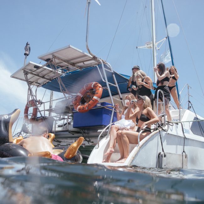 A group of people in swimwear are lounging and swimming around a boat. Inflatable toys are floating in the water. The scene appears to be a sunny day with clear skies, perfect for a private yacht charter on Sydney Harbour or a catamaran party.