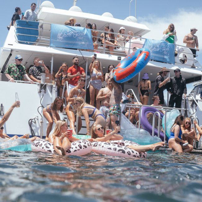 A large group of people is partying on a private yacht charter in Sydney Harbour with inflatable pool toys. Some are sitting or standing on the boat's deck, while others are in the water near the boat.