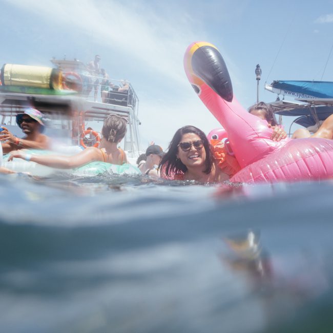 People enjoying a boat party in the water with inflatable toys and floats, including a large pink flamingo. Some individuals are swimming while others are near or on the boats. The event features DJ boat hire Sydney, making it an unforgettable experience on a sunny day.