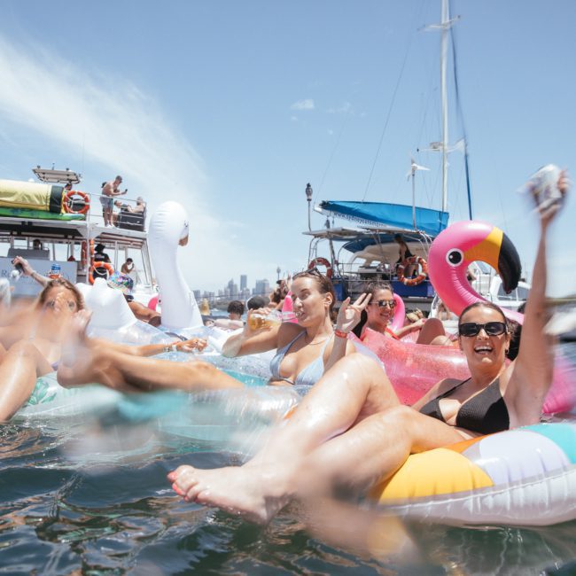 People are floating on inflatable pool toys in the water near boats on a sunny day; some are holding drinks and smiling while others gather in the background, enjoying a Sydney boat party hire.