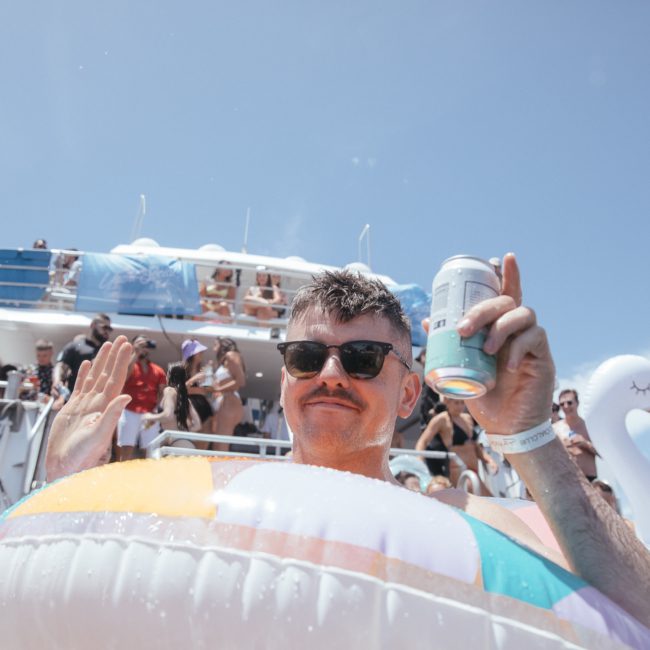 A man with sunglasses and a mustache holds a can of drink, floating on a colorful inflatable ring in the water, with a swan float nearby and a Catamaran party Sydney boat filled with people in the background.