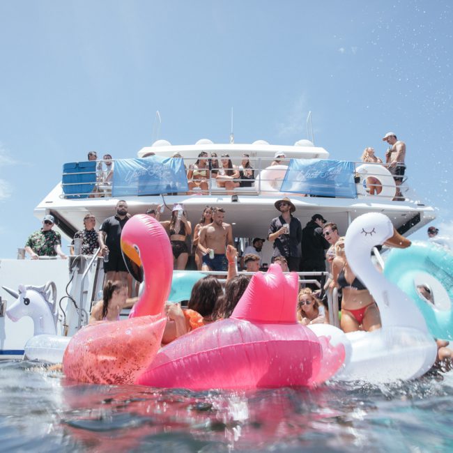 People on a private yacht charter in Sydney Harbour are partying while others are in the water with large inflatable floats, including swans and flamingos, on a sunny day.