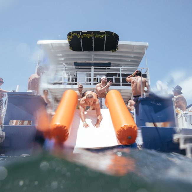 People on a luxury yacht hire in Sydney enjoying water activities, with one person sliding down an inflatable slide into the water.