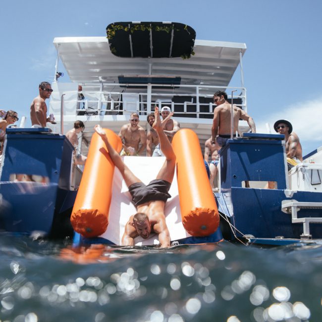 A group of people in swimsuits are on a boat. One person slides down an inflatable slide into the water while others watch, enjoying their Sydney boat party hire.