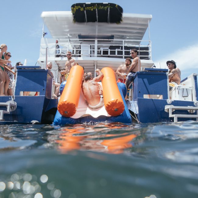A group of people relaxes on the deck of a boat while one person slides into the water on an inflatable orange slide. Clear blue sky and water surround the scene, perfect for a Sydney boat party hire.