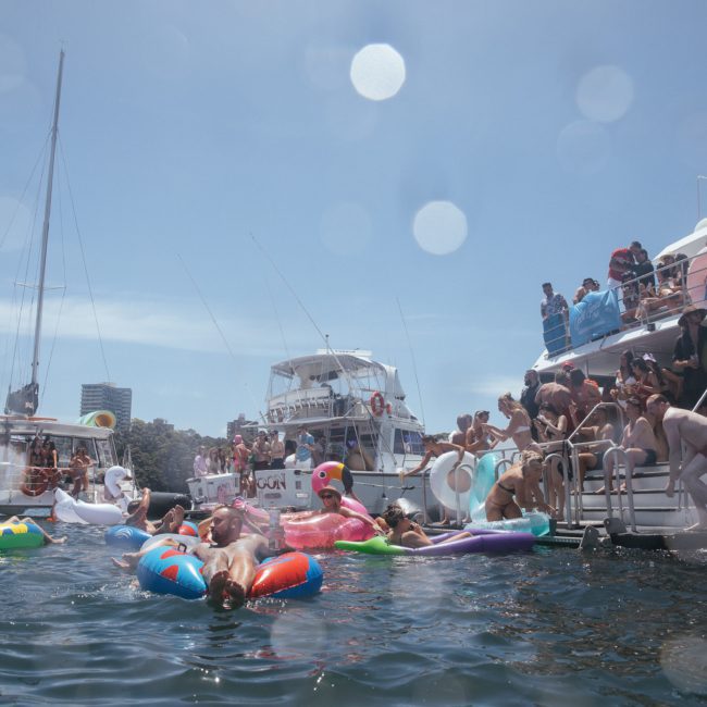 People are enjoying a sunny day on the water, swimming with colorful inflatables and gathering on docked boats in a busy, festive atmosphere. Many are taking advantage of Sydney boat party hire to elevate the celebration.