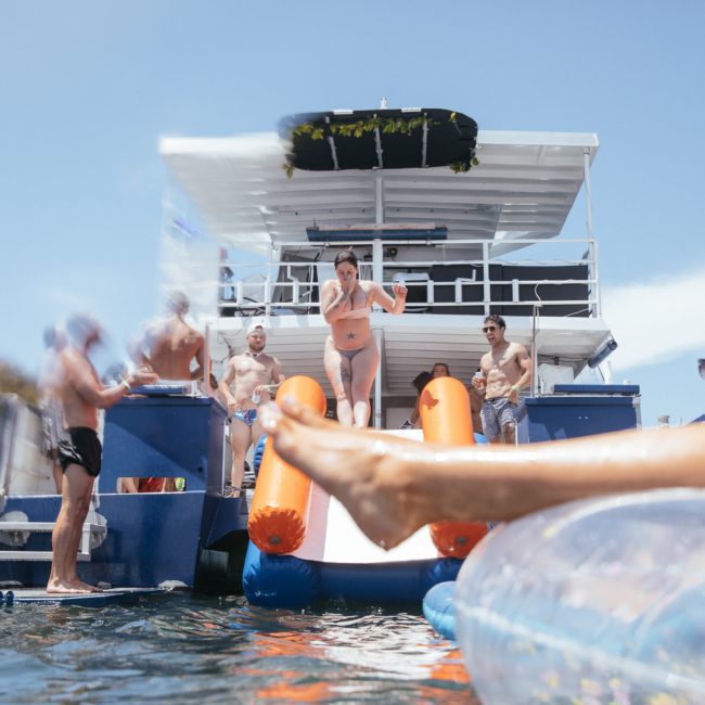 People on a private yacht charter in Sydney Harbour enjoying a sunny day; some are standing, and a woman is at the top of a water slide. An inflated raft and a person's legs are visible in the water, making it the perfect catamaran party in Sydney.