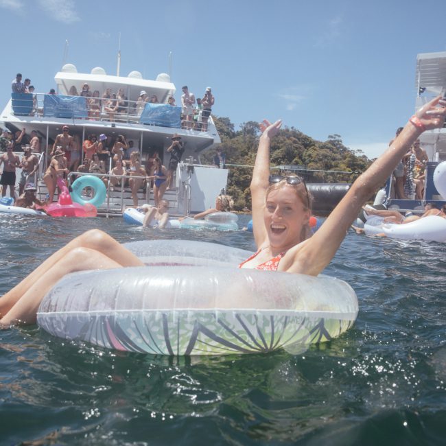 Person smiling in a pool float raises arms in the water, with two boats and several people enjoying the Sydney boat party hire experience in the background.