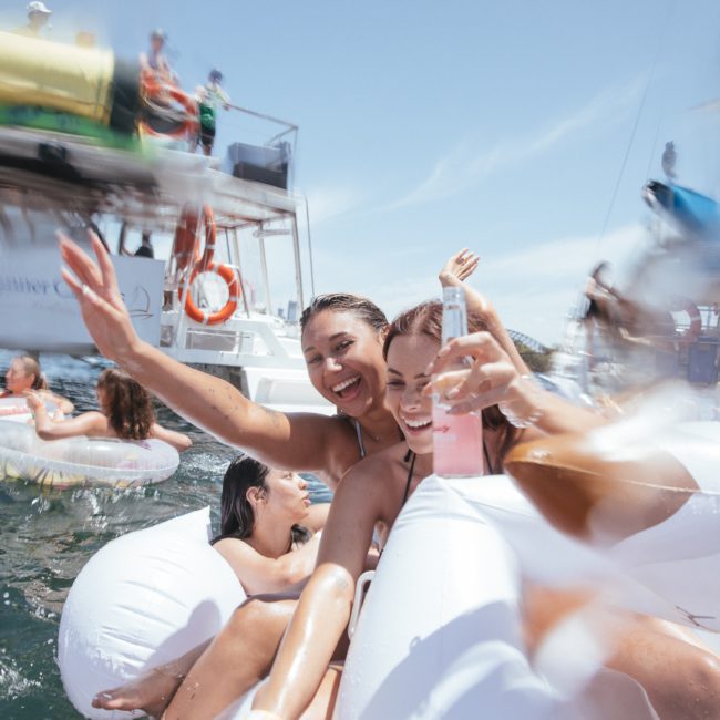 Two smiling women in swimsuits are enjoying a sunny day on the water, holding drinks while floating on inflatable tubes during a Sydney boat party hire, with boats and other people around them.