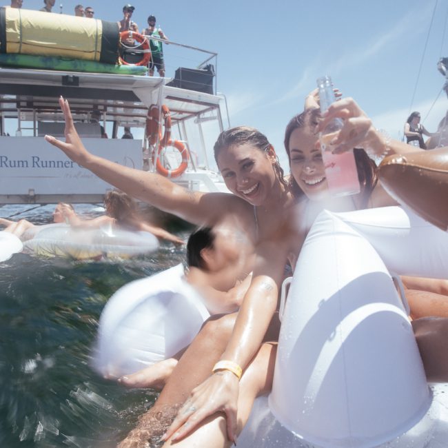 Two women on inflatable floaties in the water pose for a photo, smiling and holding drinks. A boat labeled 'Rum Runner' and more people are seen in the background, enjoying their time. Perfect setting for a Sydney boat party hire!