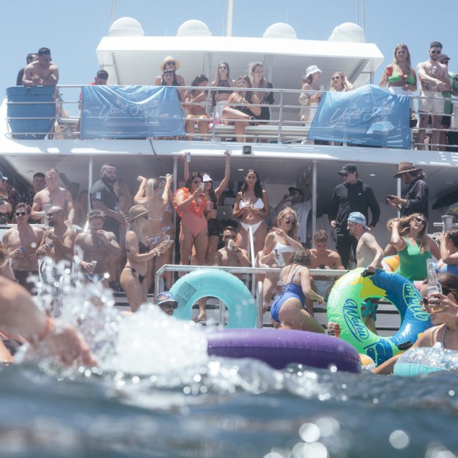 A group of people on a boat and in water, some standing, others using inflatables. The back of the boat has a banner for DJ boat hire Sydney and people are enjoying a sunny day, taking photos and relaxing.