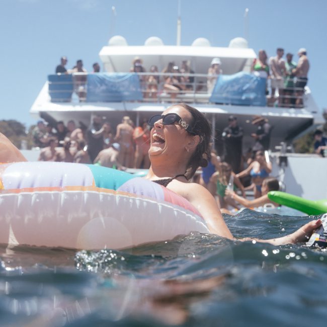 A woman relaxes on a colorful float in the water, holding a drink, with a luxury yacht hire Sydney in the background during a sunny day.