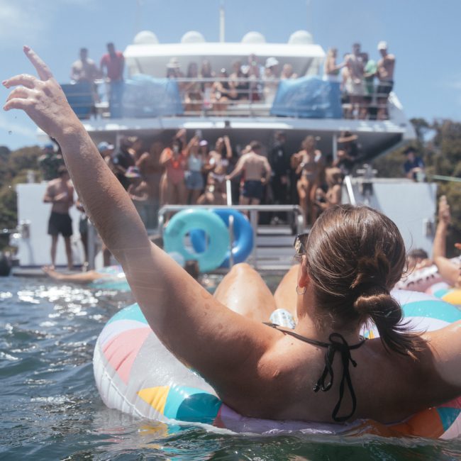 A person on a colorful inflatable floats in water, arms raised, as a crowd socializes on a nearby private yacht charter in Sydney Harbour with multiple decks. The boat is moored near a green, forested shoreline.