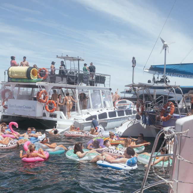 People relax on inflatable floats and boats in the water near a docked catamaran on a sunny day. Some participants are on board the boats while others are floating in the water, enjoying what feels like the perfect catamaran party Sydney has to offer.