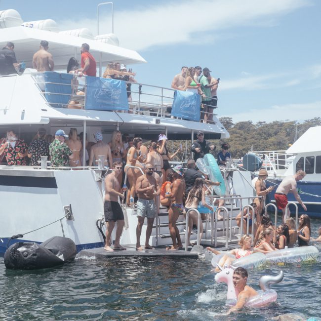 A group of people enjoying a sunny day on a luxury yacht, with some swimming in the water using inflatables, and others mingling on the boat's deck. Another yacht is visible on the right side of the image, showcasing an ideal setting for a Sydney boat party hire.