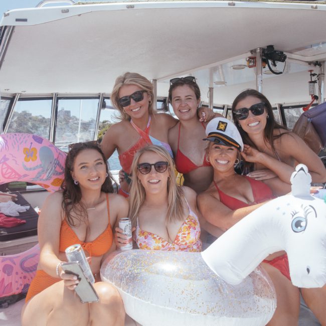 A group of six women in swimsuits poses on a boat, holding drinks and inflatable floaties, with sunlit water and other boats in the background, enjoying a lively Sydney boat party hire.