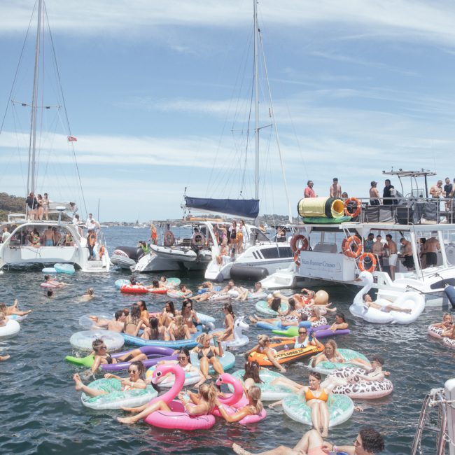 People are enjoying a sunny day on a large body of water, lounging on colorful inflatable floats. Boats, including yachts and sailboats, are anchored nearby—some even indulging in DJ boat hire Sydney services for added fun.