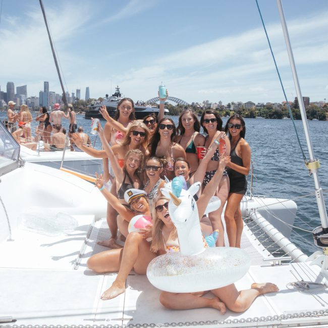 A group of people in swimwear pose on a private yacht charter in Sydney Harbour, some with drinks, in front of a cityscape and bridges over water on a sunny day. An inflatable unicorn float is in the foreground.