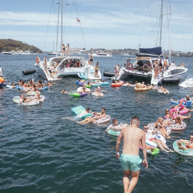 People are enjoying a sunny day on a lake with boats and inflatables, relaxing and swimming in the water. The scene could easily be part of a catamaran party in Sydney, adding a touch of excitement to the day.