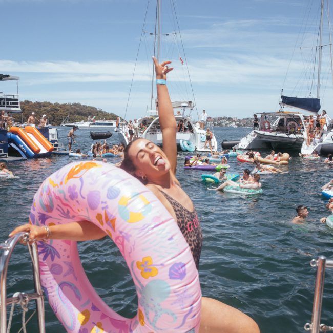 A woman wearing a pink inflatable ring poses jubilantly on a boat at a lively Sydney boat party hire, surrounded by people swimming and floating in the water on a sunny day. Several anchored boats and inflatable toys are in the background, capturing the essence of fun during this private yacht charter Sydney Harbour event.