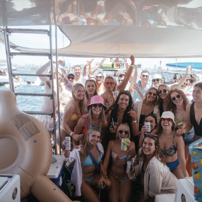 A large group of people in swimwear are gathered on a luxury yacht, smiling and posing for the camera. The backdrop features more boats and Sydney Harbour under a sunny, clear sky.