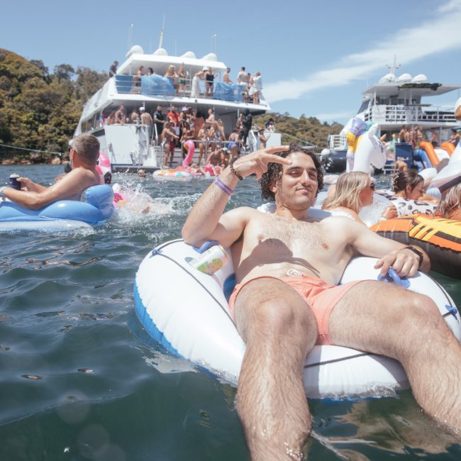 A man reclines on an inflatable pool float in the water, surrounded by other people on floats and boats, enjoying a sunny day.