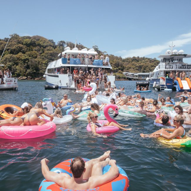 A crowd of people in colorful inflatable floats enjoy a sunny day on the water near anchored yachts, perfect for a Sydney boat party hire.
